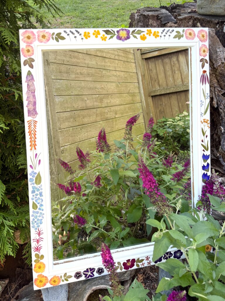 A pressed flower decorated mirror frame surrounded by blooming garden plants